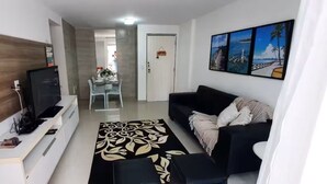 A modern living room with a flat-screen TV, a black sofa, a coffee table, and a dining area with a glass table and chairs.