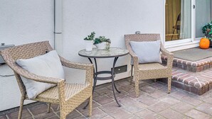 Fletstol med hynder, et lille rundt bord og potteplanter på en terrasse med en hvid væg og murstens trapper.