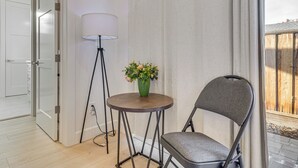 Et hjørneværelse med en stol, et lille bord med en blomstervase, en gulvlampe og udsigt til et træhegn udenfor.