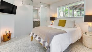 A modern bedroom with a large bed, a TV mounted on the wall, a bedside table with a lamp, and a window with blinds. - Tropical Backyard W/pool: 10 min From Miami Beach! (Miami)