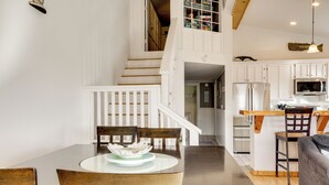 A dining area with a round table, chairs, and a decorative centerpiece. A staircase leads to a lofted space with a bookshelf. - Stroll to Shore: Retreat w/ Balcony in Gold Beach (Gold Beach)