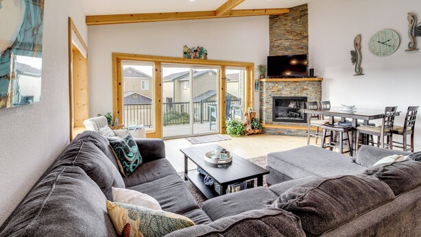 A spacious living room with a large gray sofa, a coffee table, and a fireplace. There is a dining area with a table and chairs, and a balcony with sliding glass doors. - Stroll to Shore: Retreat w/ Balcony in Gold Beach (Gold Beach)