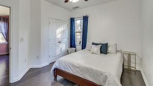Ein Schlafzimmer mit einem Bett, einem Deckenventilator, einem Fenster mit Vorhängen und einem kleinen Tisch.