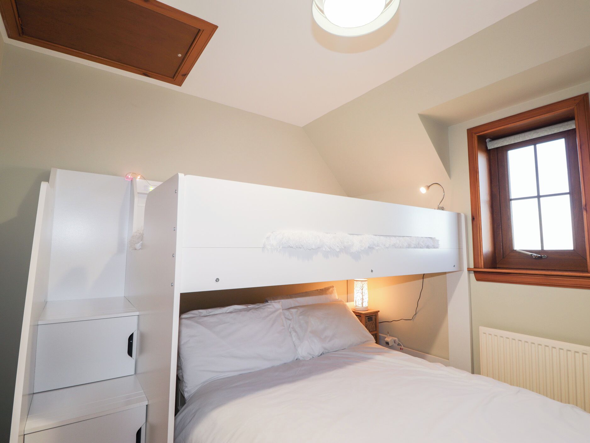 A bedroom with a bunk bed, a bedside table, a lamp, and a window with a view of the outside.