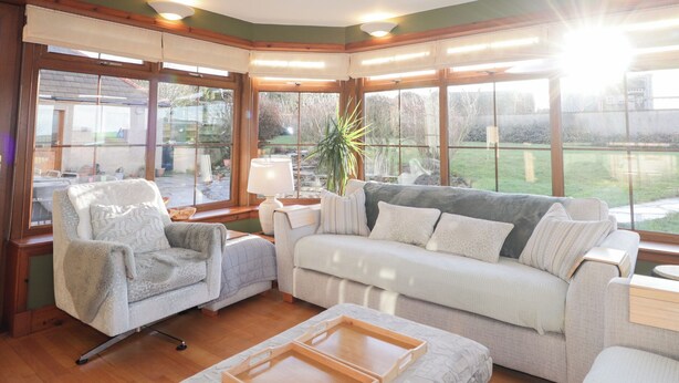 A cozy living room with a sofa, armchair, and a coffee table. Large windows offer a view of the backyard. - Northbanks House (Thurso)