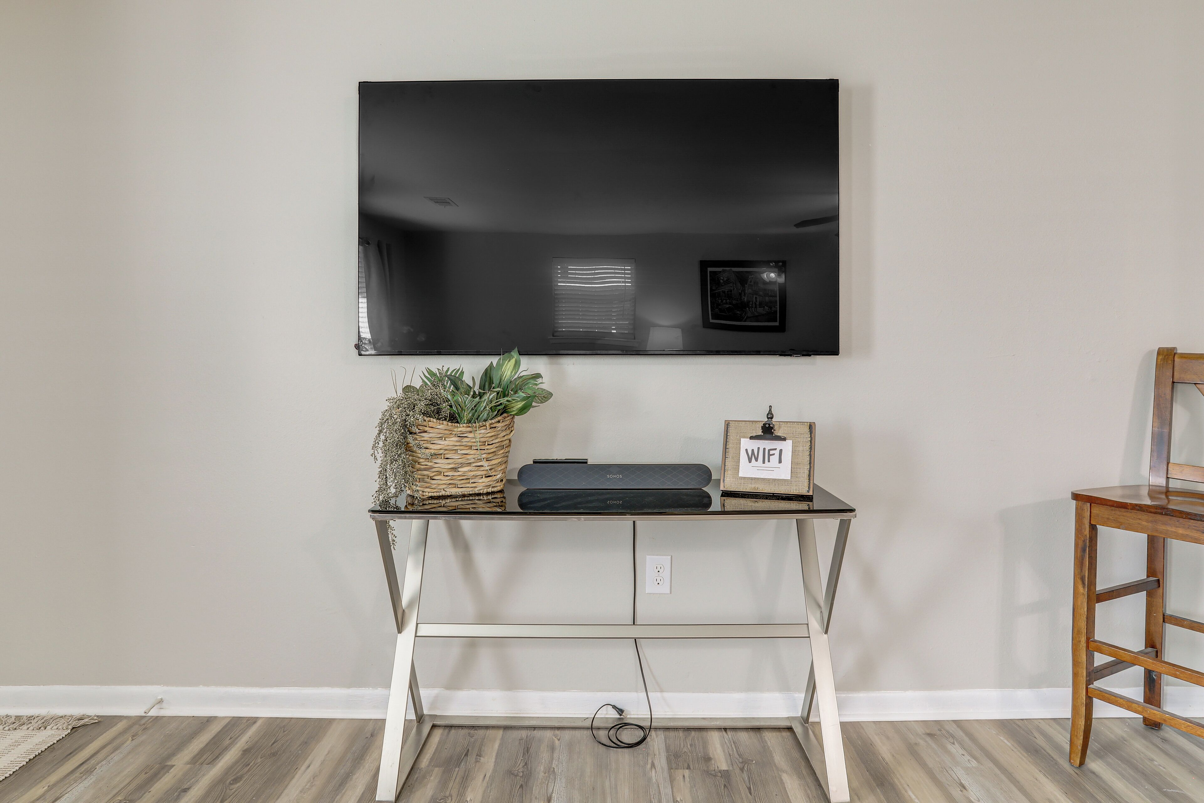 A modern living room with a flat-screen TV mounted on the wall, a small wooden side table with a plant and a framed picture, and a white console table with a power strip.