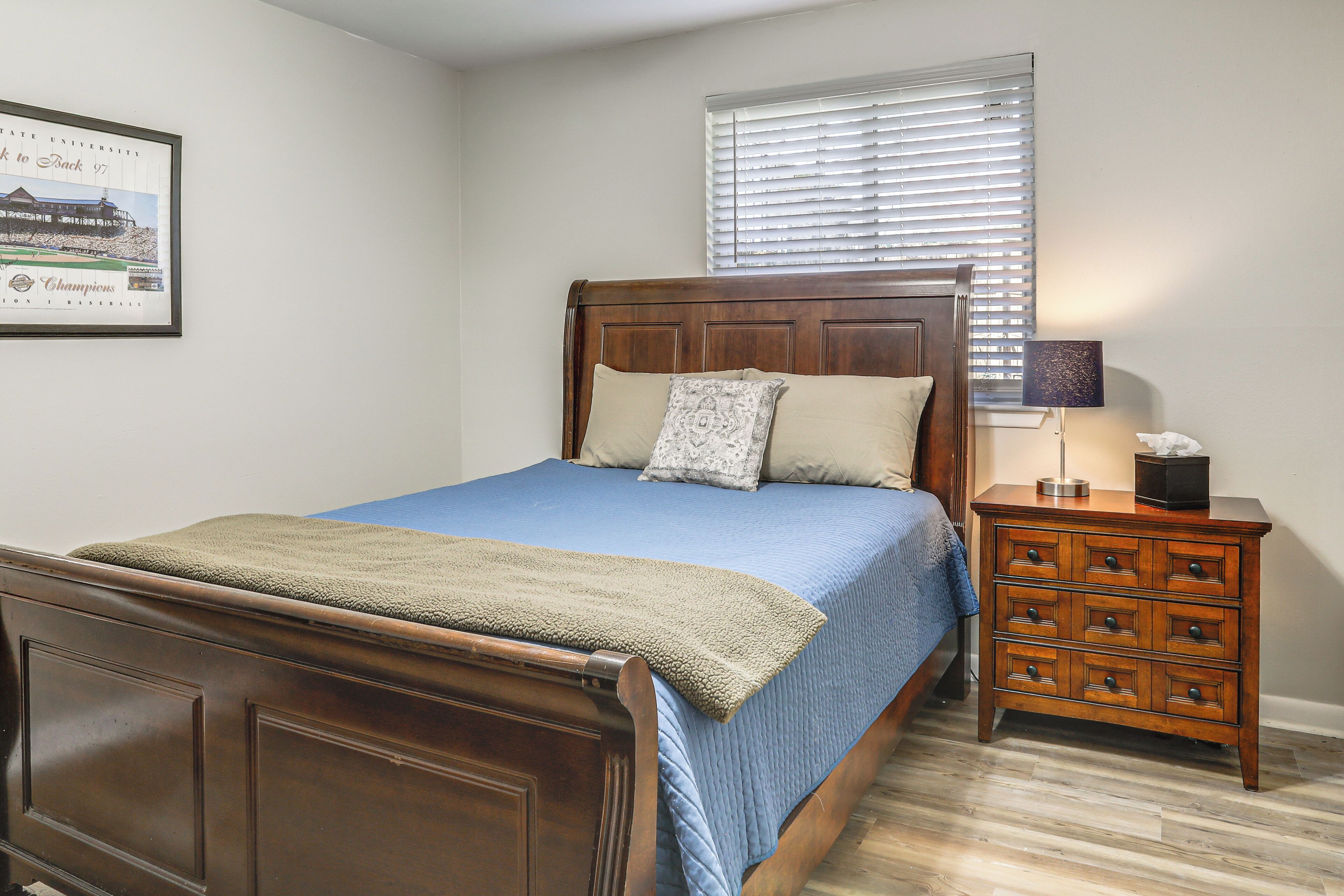 Un dormitorio con cama de madera, colcha azul, mesita de noche con cajones, un cuadro enmarcado en la pared y una ventana con persianas.