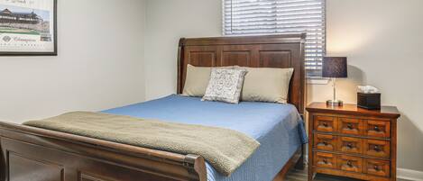 Un dormitorio con cama de madera, colcha azul, mesita de noche con cajones, un cuadro enmarcado en la pared y una ventana con persianas.