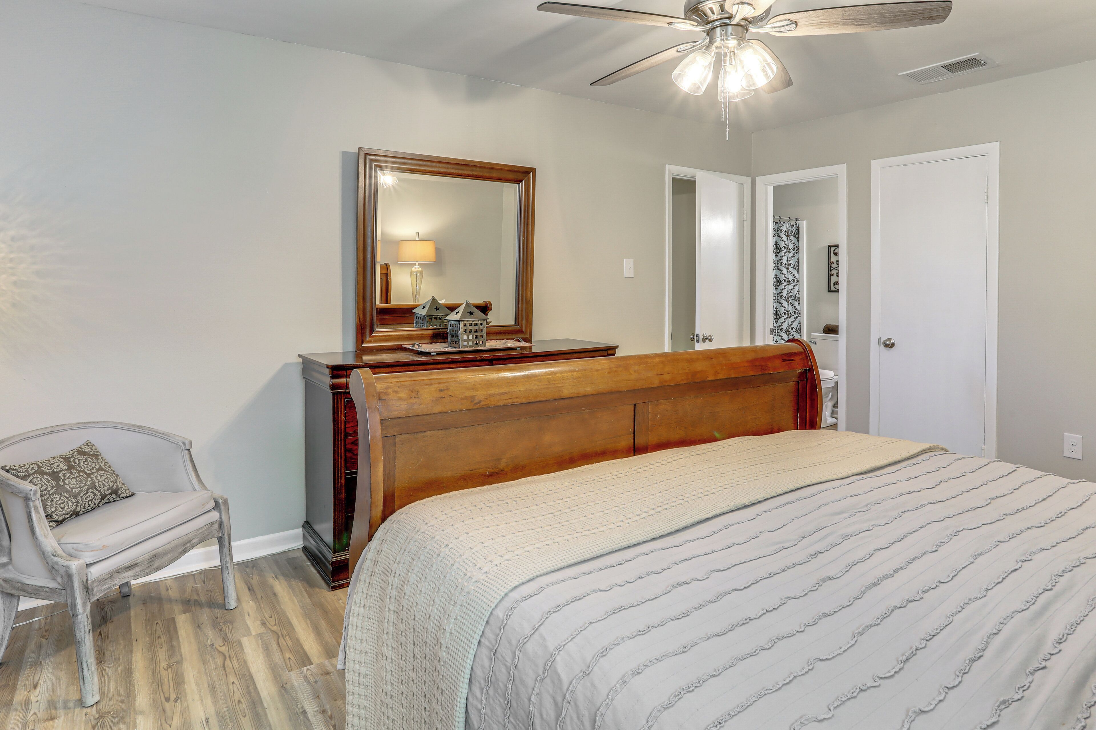 A bedroom with a wooden headboard, a large bed with a striped bedspread, a wooden nightstand, a chair, a mirror, and a ceiling fan.