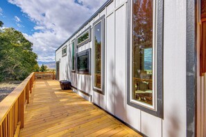 Una terraza de madera con barandilla, ventanas grandes y vistas a árboles y montañas.