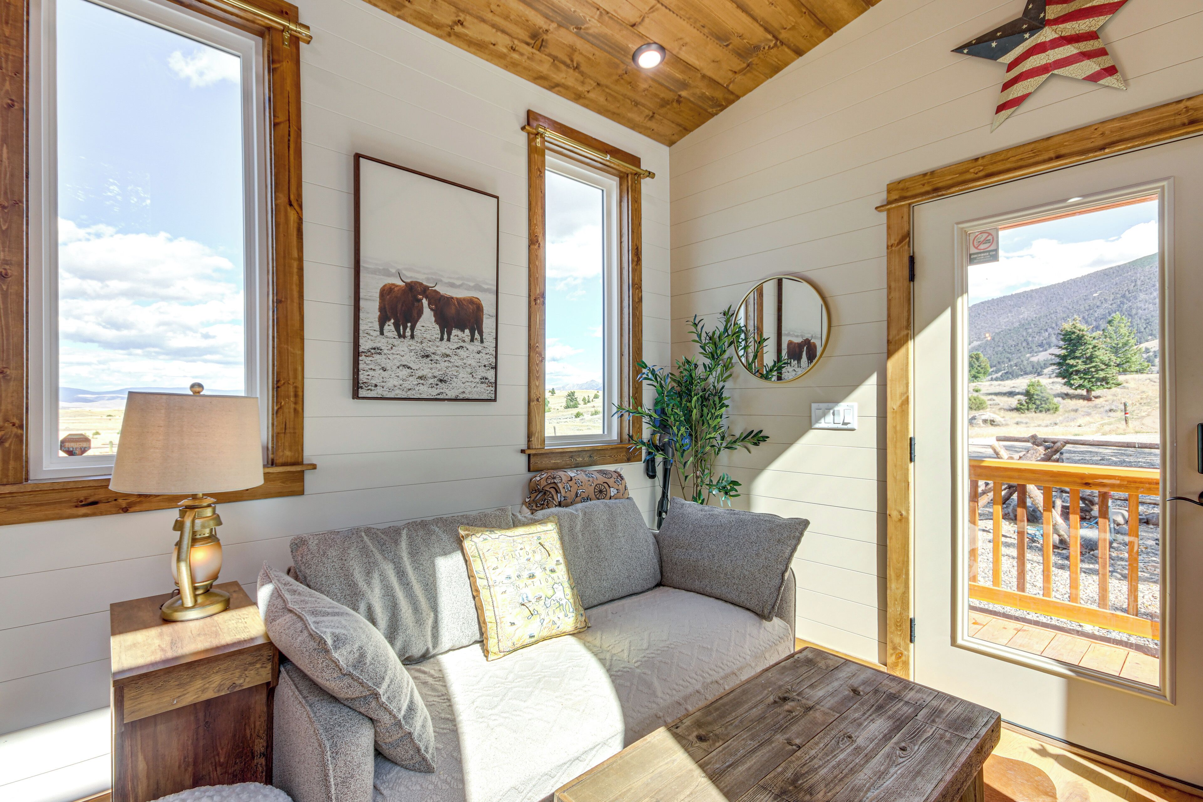 A cozy room with a wooden ceiling, a sofa with cushions, a table, a lamp, a framed picture of a bull, a mirror, and a door leading to an outdoor deck.