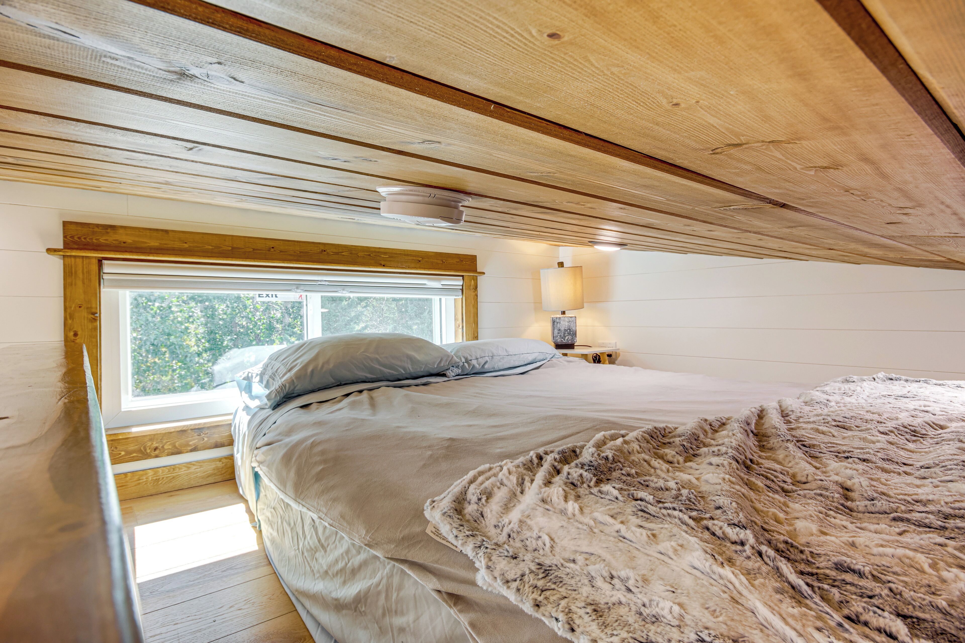 A bedroom with a large window, wooden ceiling beams, a bed with a fluffy blanket, and a nightstand with a lamp.