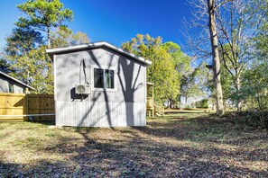 Une petite cabane à un seul étage, avec un toit en pente, une fenêtre et une porte. Elle est entourée d’une clôture en bois et d’arbres.