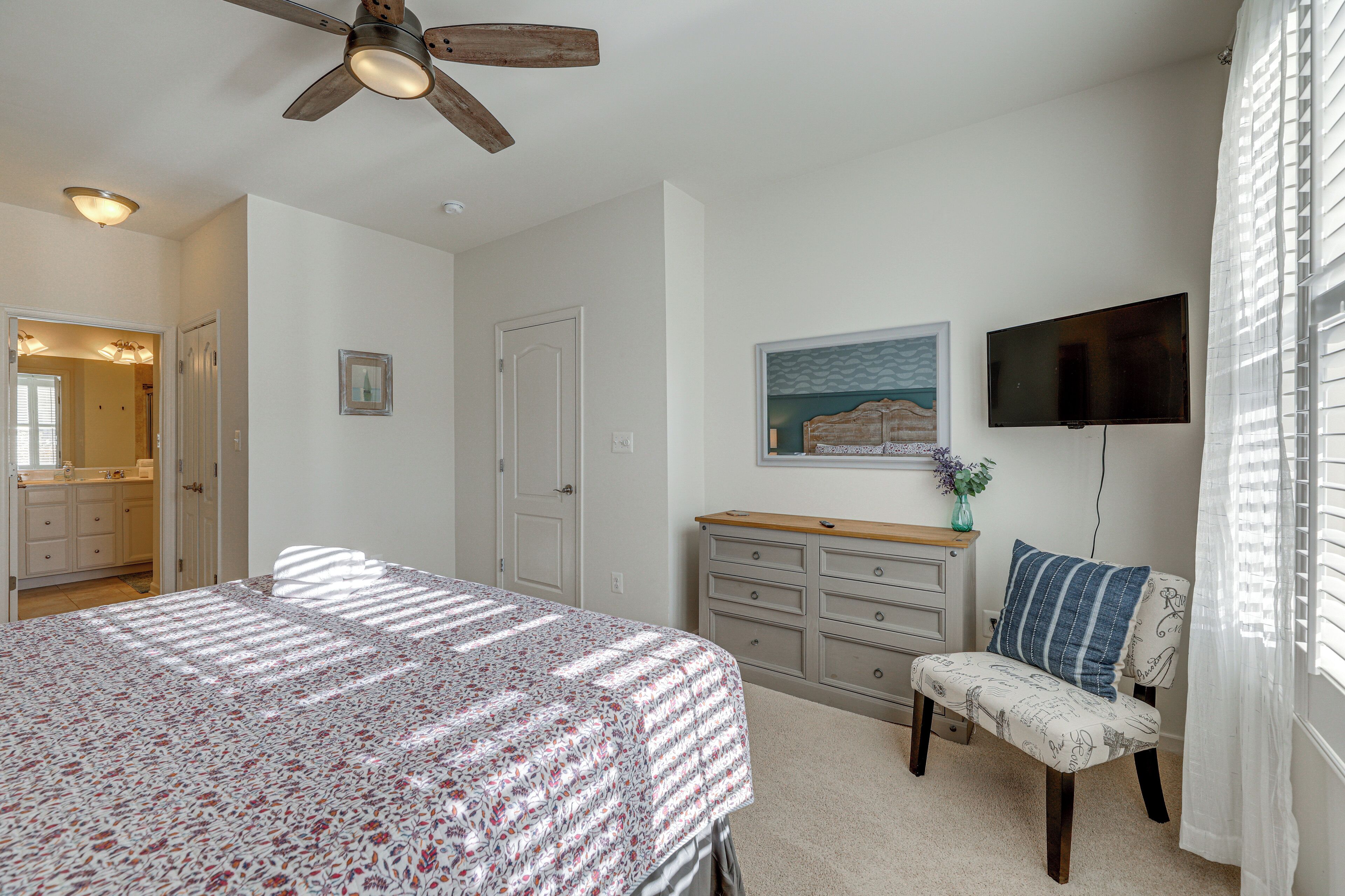 A bedroom with a bed, a dresser, a chair, a ceiling fan, a TV, and a painting on the wall.