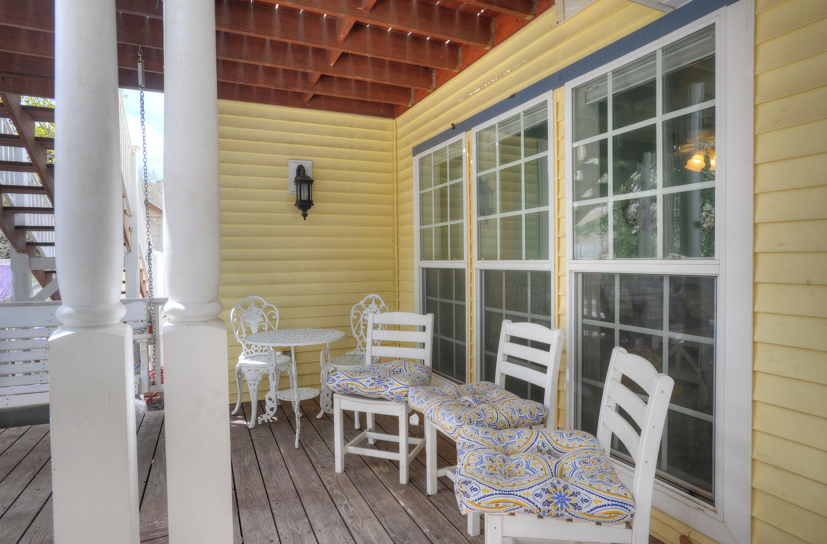 Une véranda couverte avec des chaises blanches et une table, un plafond en bois et un mur jaune.