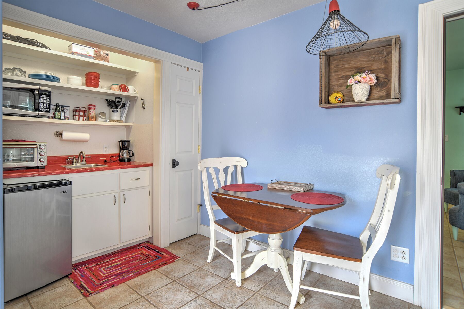 A compact kitchen with white cabinetry, a red countertop, a small dining table with two chairs, a red hanging light, and a wooden shelf with a vase.