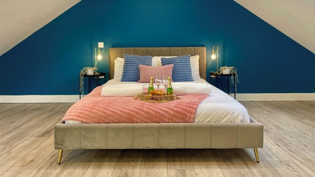 Une chambre avec un plafond en pente, un lit recouvert de plusieurs coussins, des tables de chevet avec des lampes et un plateau contenant divers objets posé sur le lit.