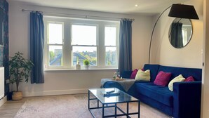 Un salon avec un canapé bleu, une table basse en verre, une plante en pot et une vue sur les immeubles à travers de grandes fenêtres.