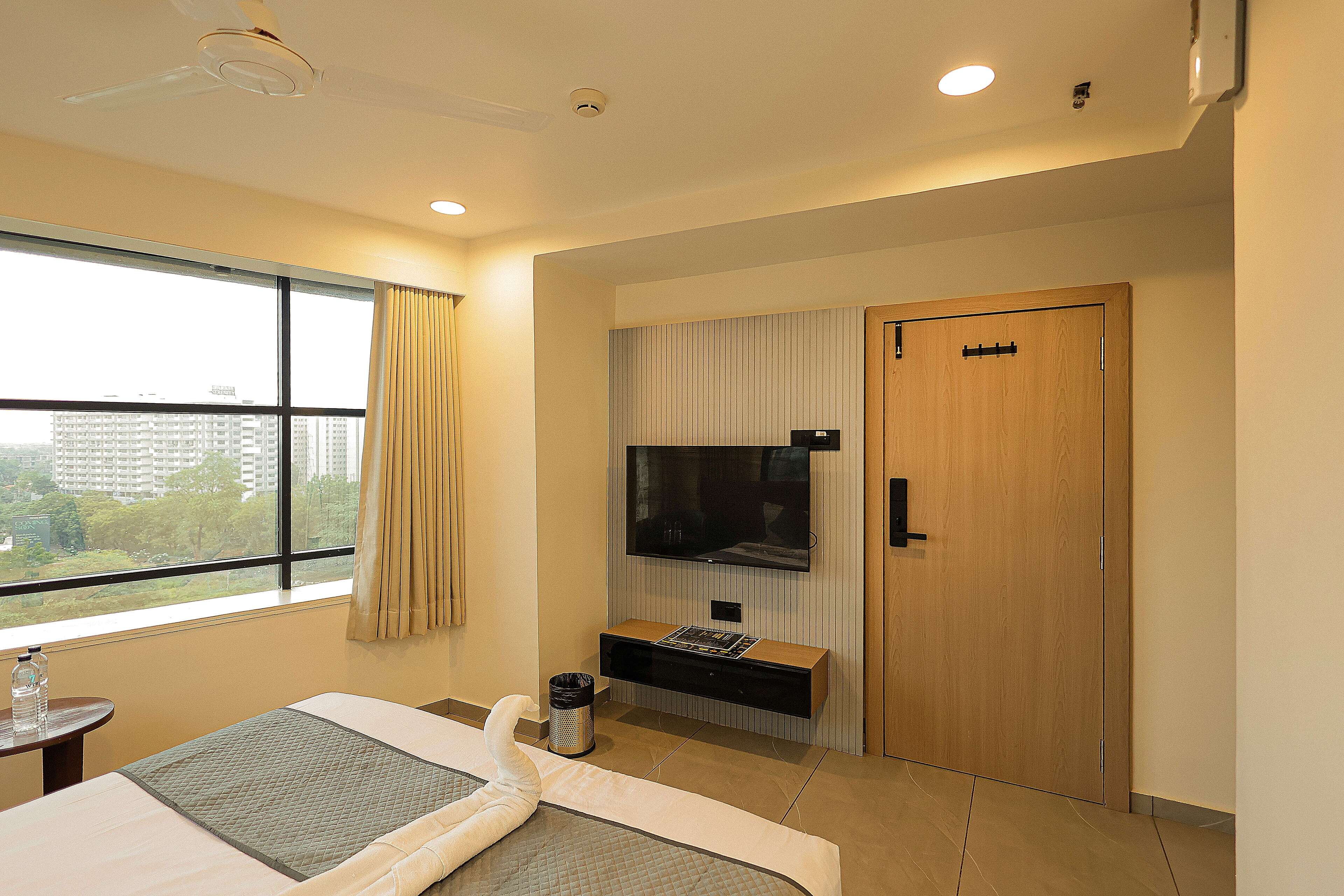 A hotel room with a bed, a TV mounted on the wall, a wooden door, and a window with curtains.