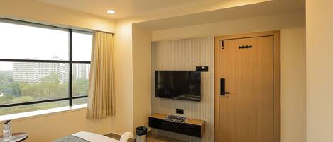 A hotel room with a bed, a TV mounted on the wall, a wooden door, and a window with curtains.
