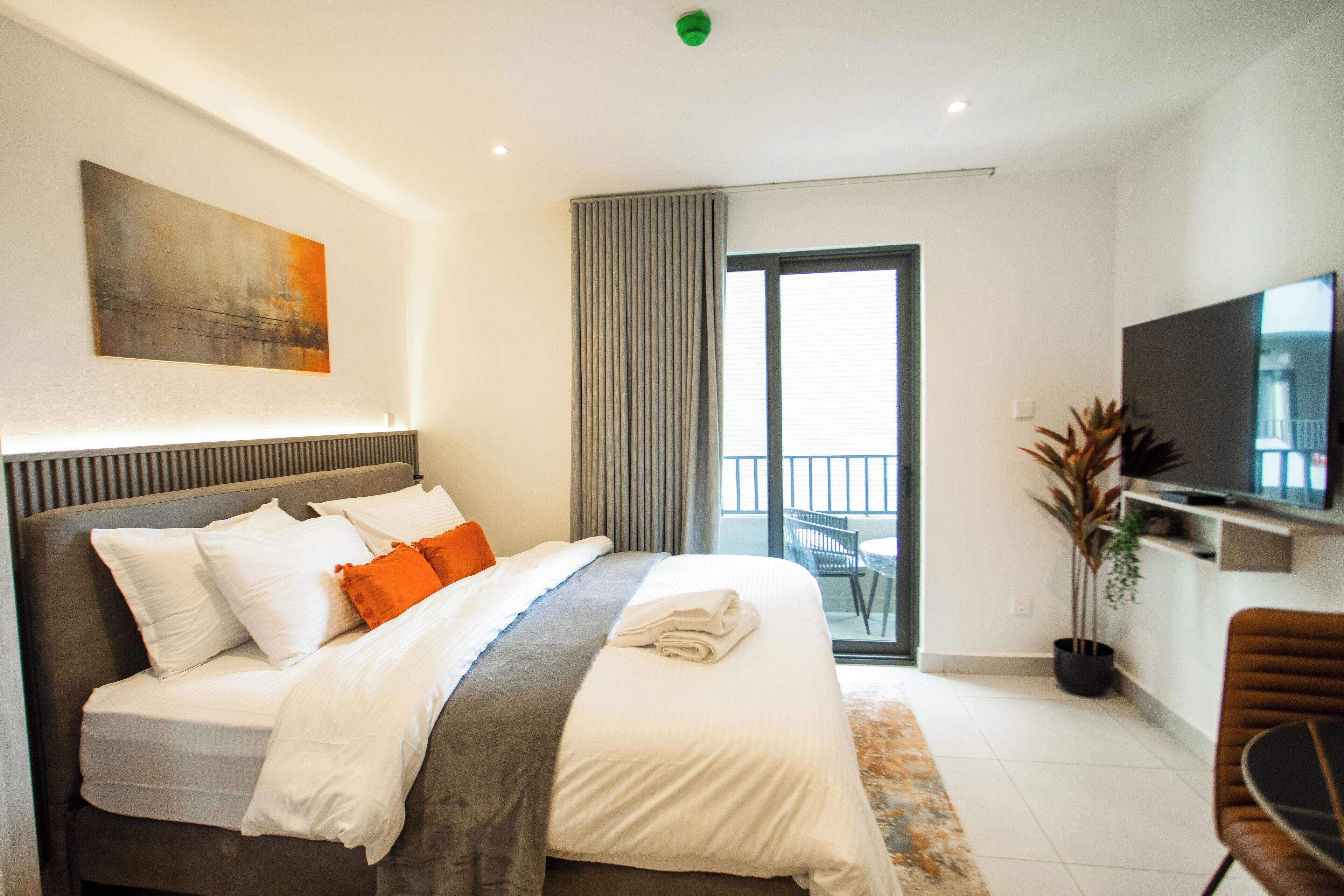 A modern hotel room with a large bed, a flat-screen TV, a balcony with a view, and a painting on the wall.