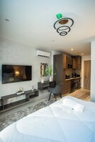 A modern hotel room with a large bed, a flat-screen TV mounted on the wall, a small seating area, and a kitchenette.