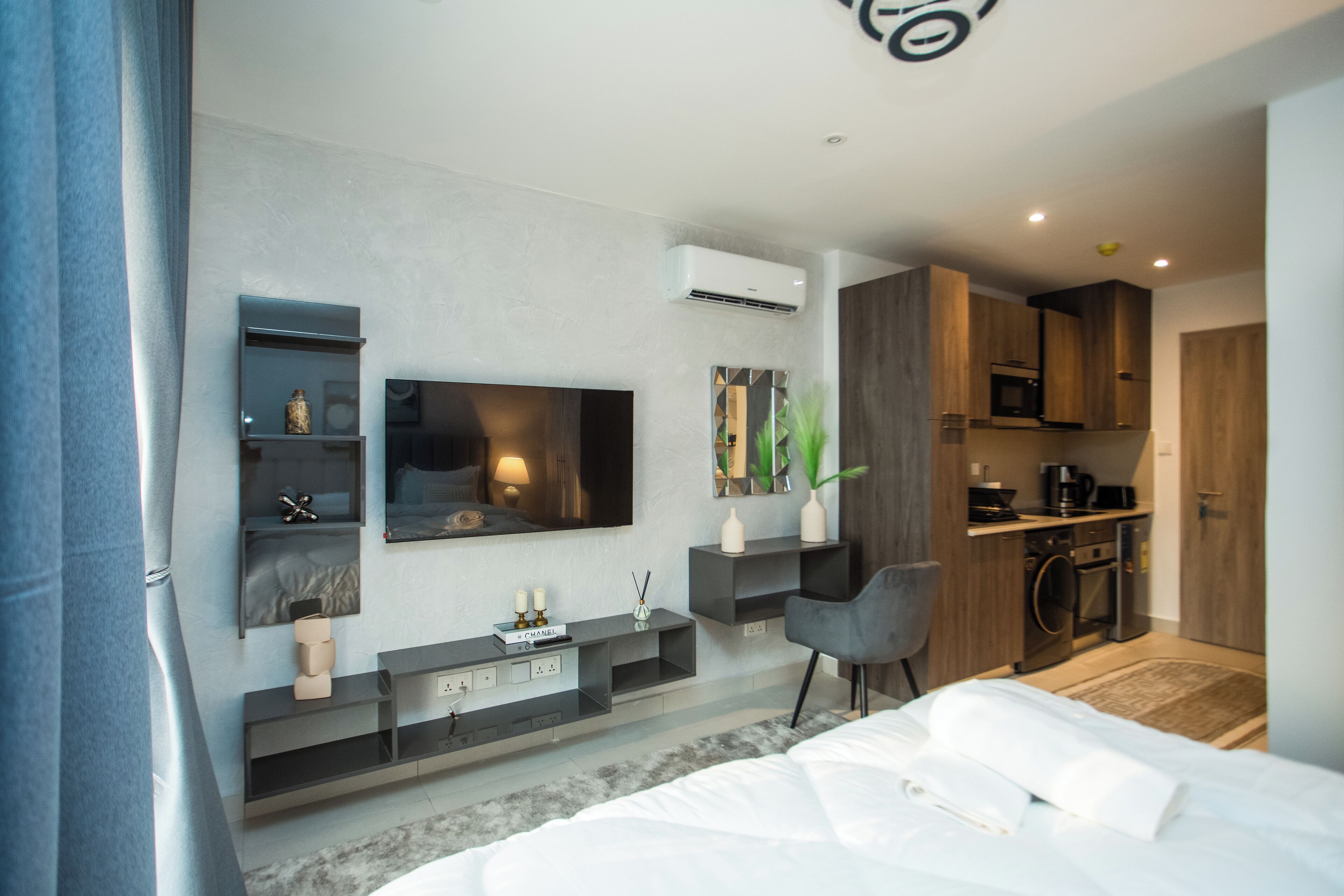 A modern hotel room with a large bed, a TV mounted on the wall, a small desk area, and a kitchenette.