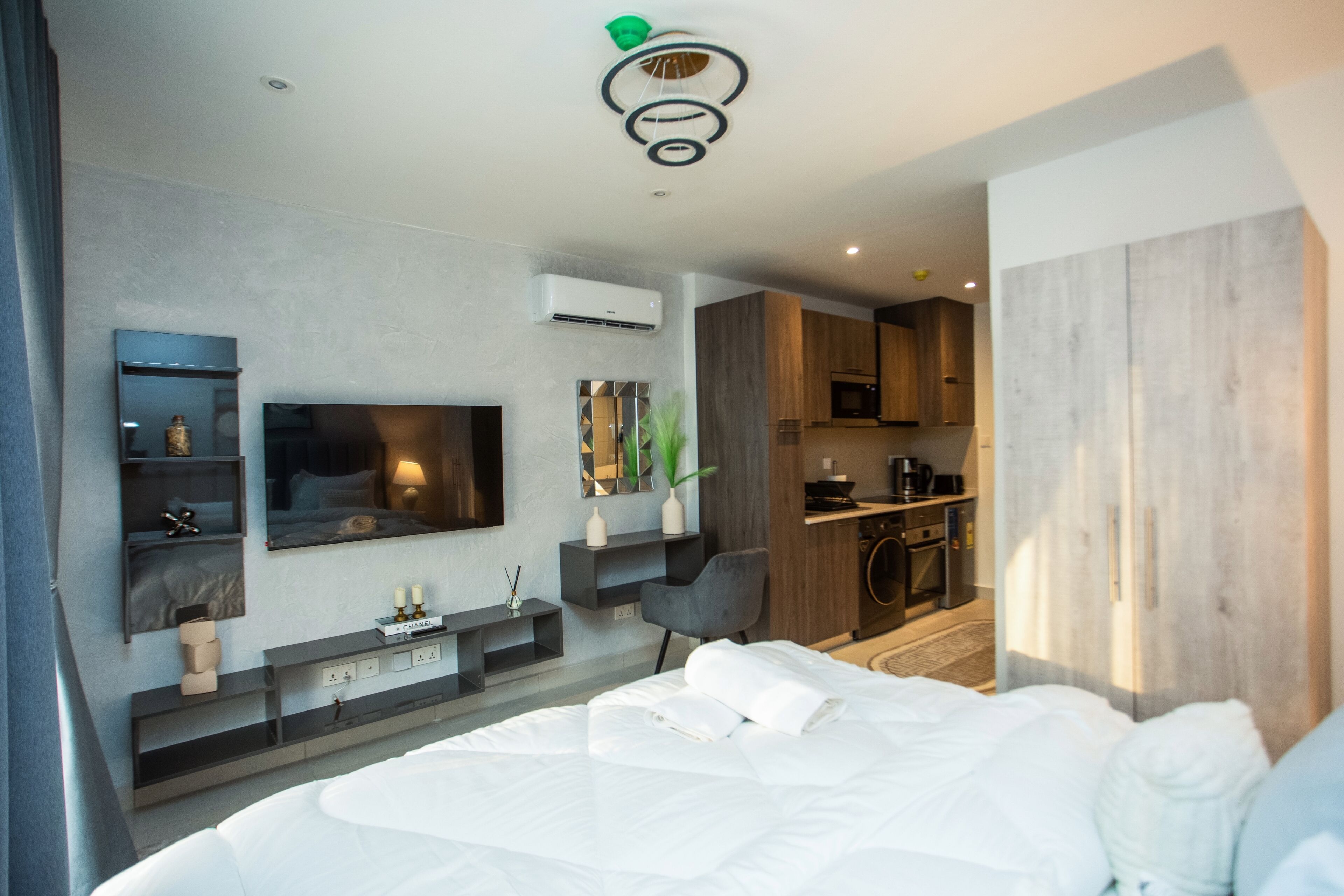 A modern hotel room with a large bed, a flat-screen TV mounted on the wall, a small seating area with a chair, and a kitchenette in the background.
