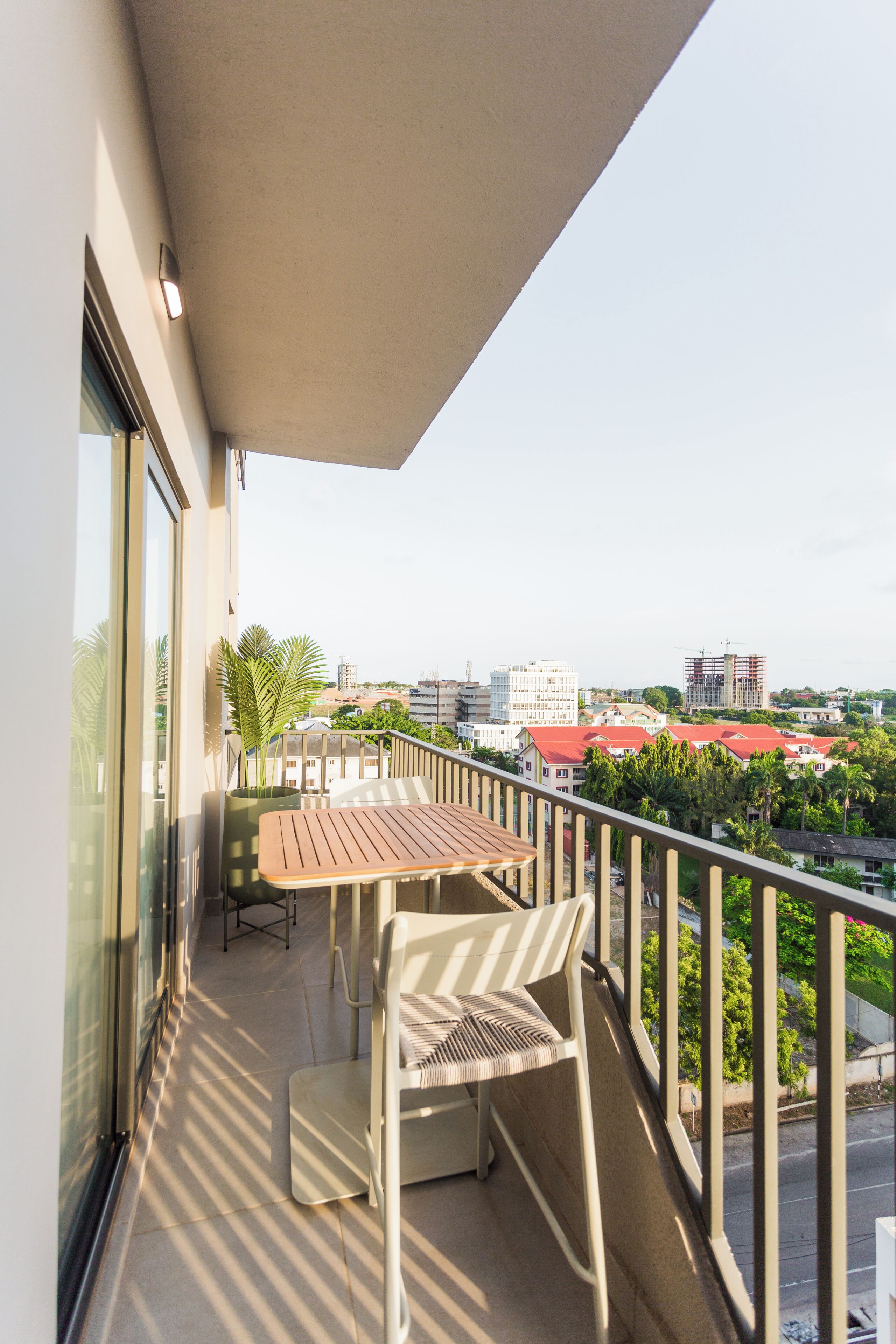 A balcony with a table and chairs, a potted plant, and a view of buildings and greenery.