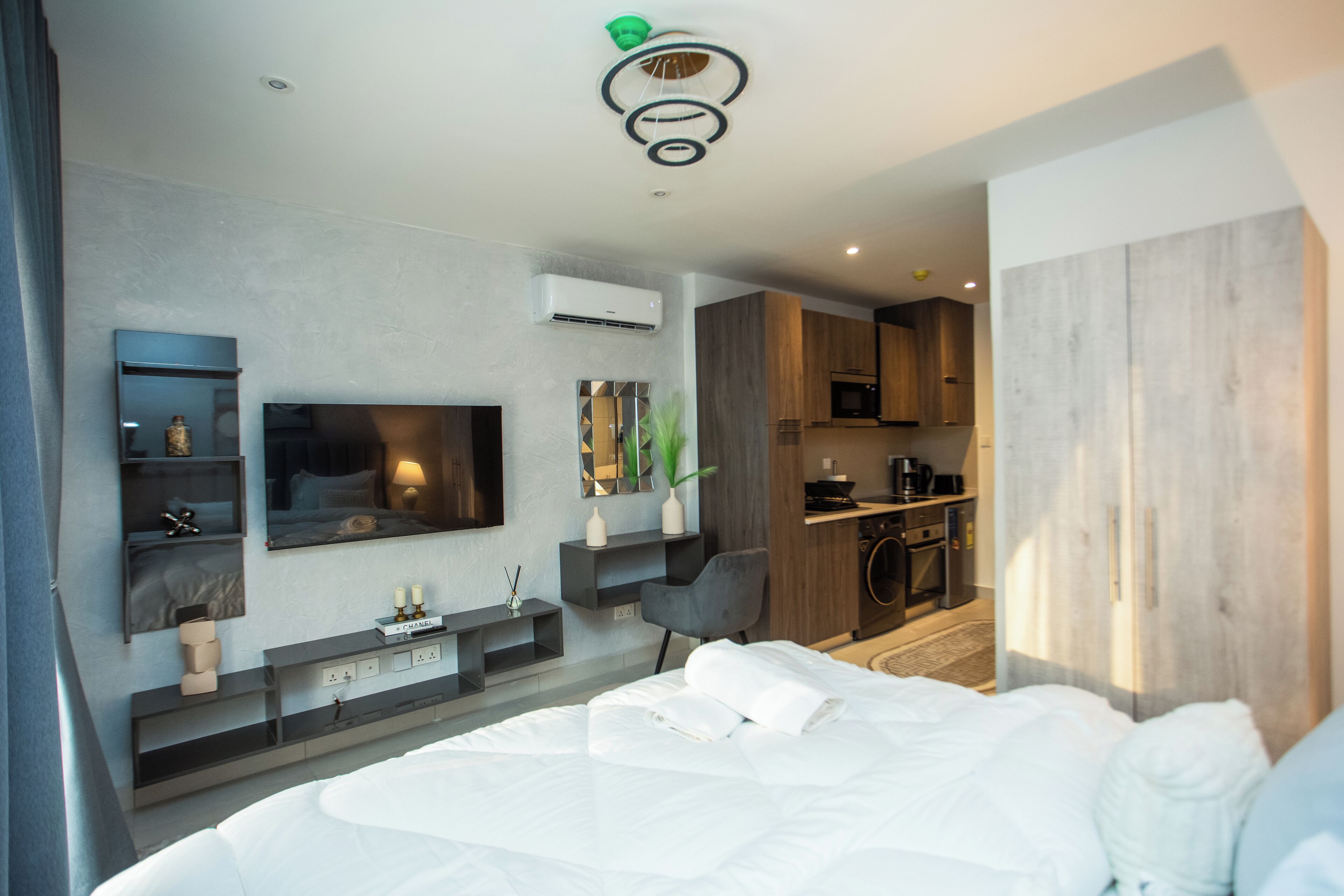 A modern hotel room with a large bed, a flat-screen TV mounted on the wall, a small seating area with a chair, and a kitchenette in the background.