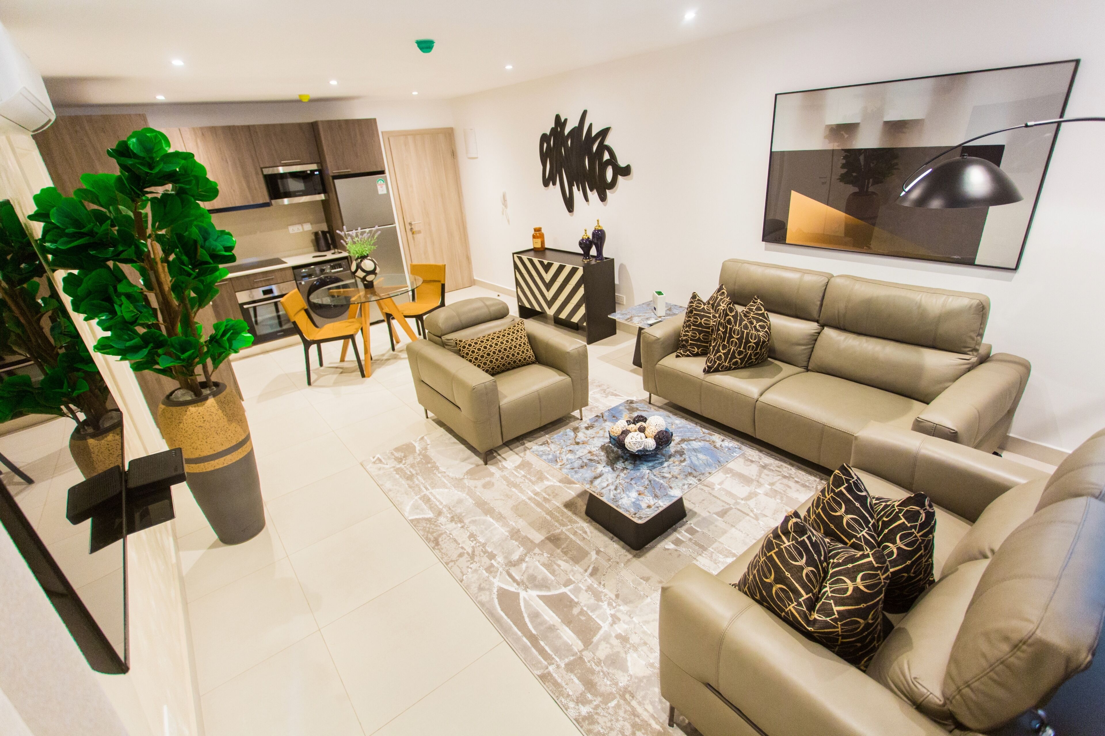 A modern living room with a sofa, coffee table, and decorative pillows. A dining area with chairs and a table is visible in the background.