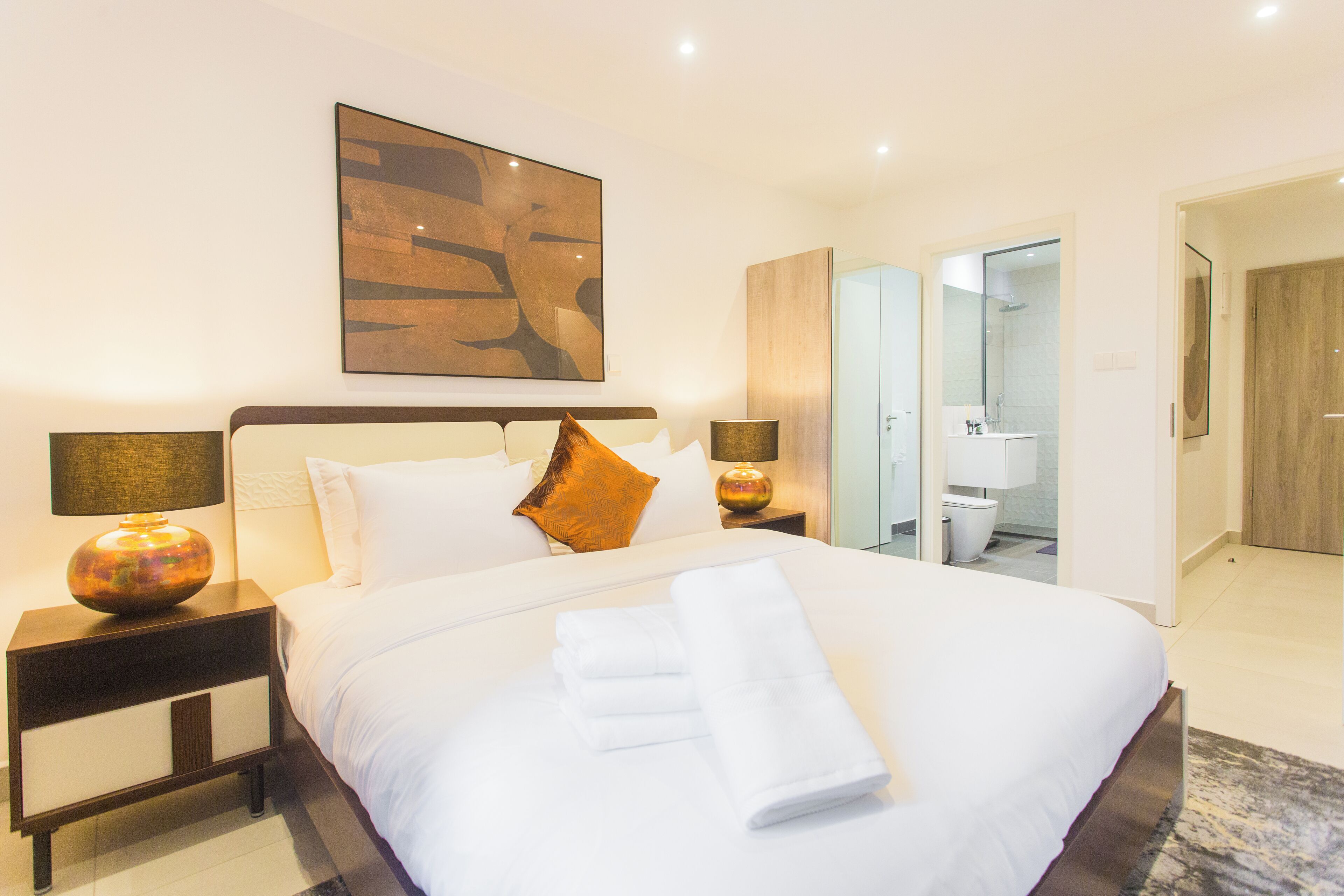 Ein Hotelzimmer mit einem großen Bett, zwei Nachttischlampen, einem Gemälde über dem Bett und einem durchgegangenen Türrahmen einsehbarer Badezimmers.