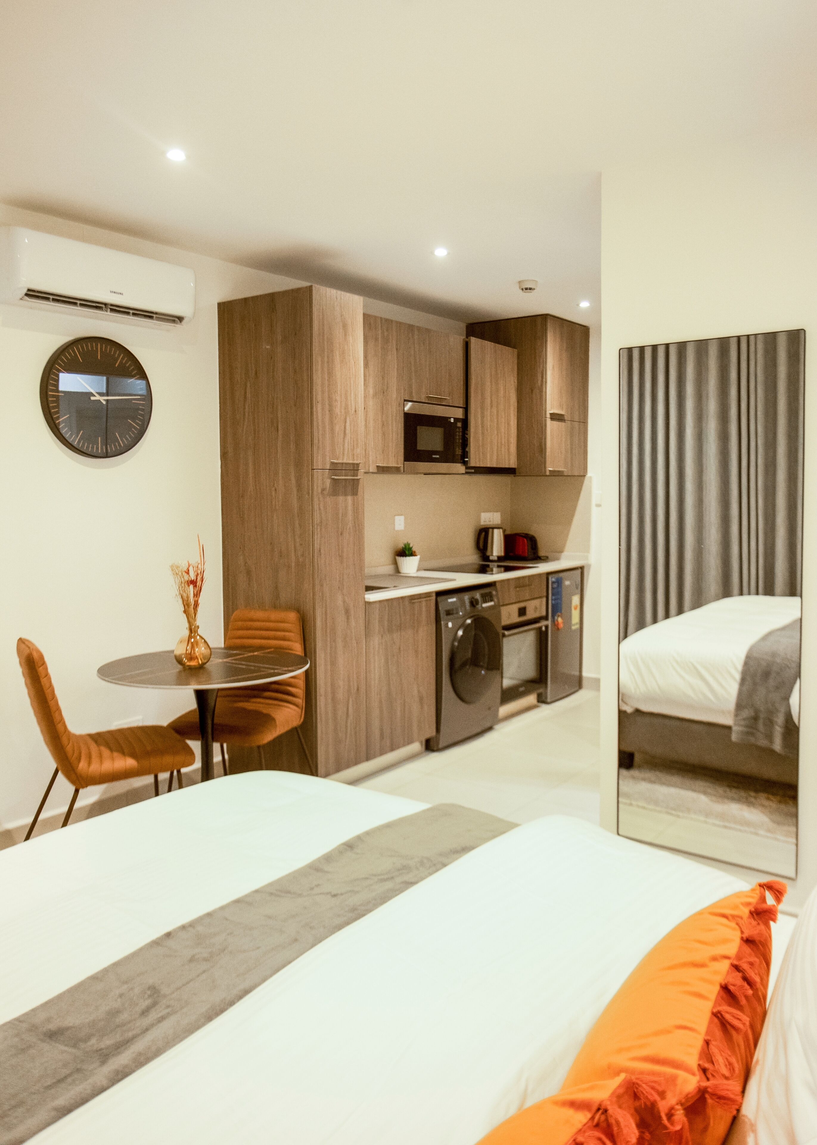 A modern hotel room with a bed, a small dining table with chairs, a kitchen area with a microwave, and a washing machine.