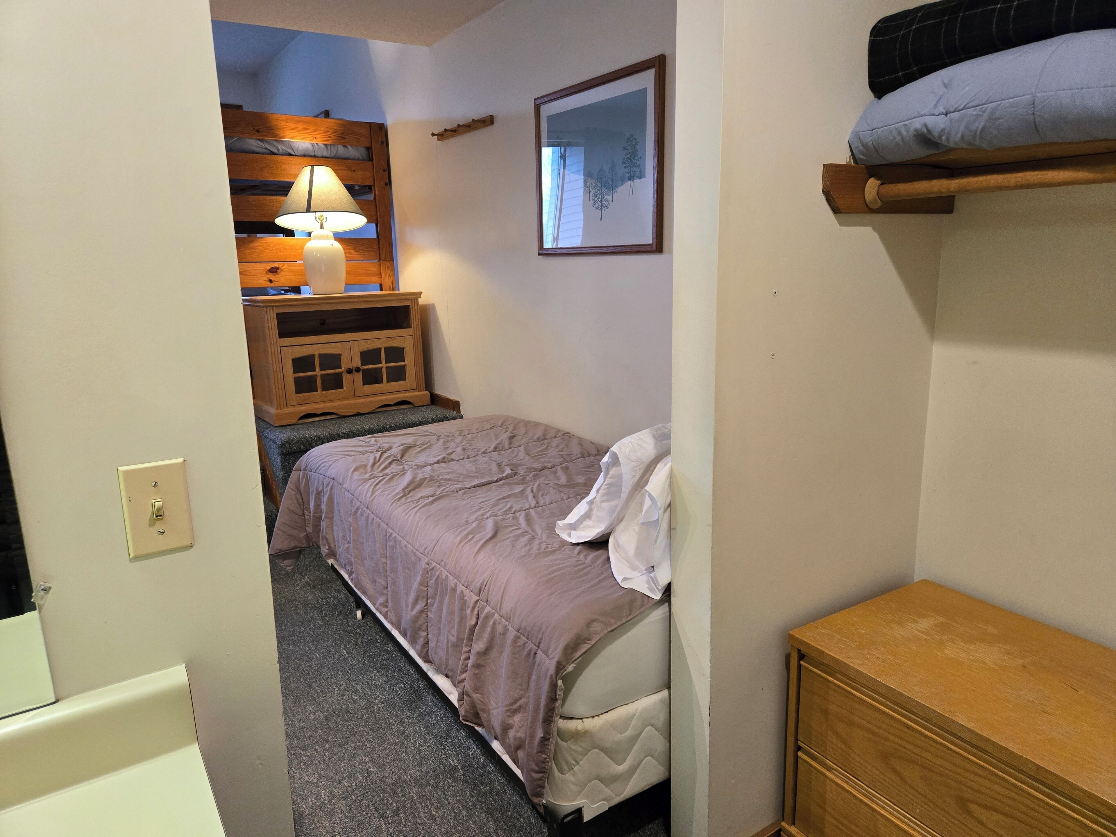 Een slaapkamer met een bed, een nachtkastje met een lamp, een dressoir en een ingelijst schilderij aan de muur.