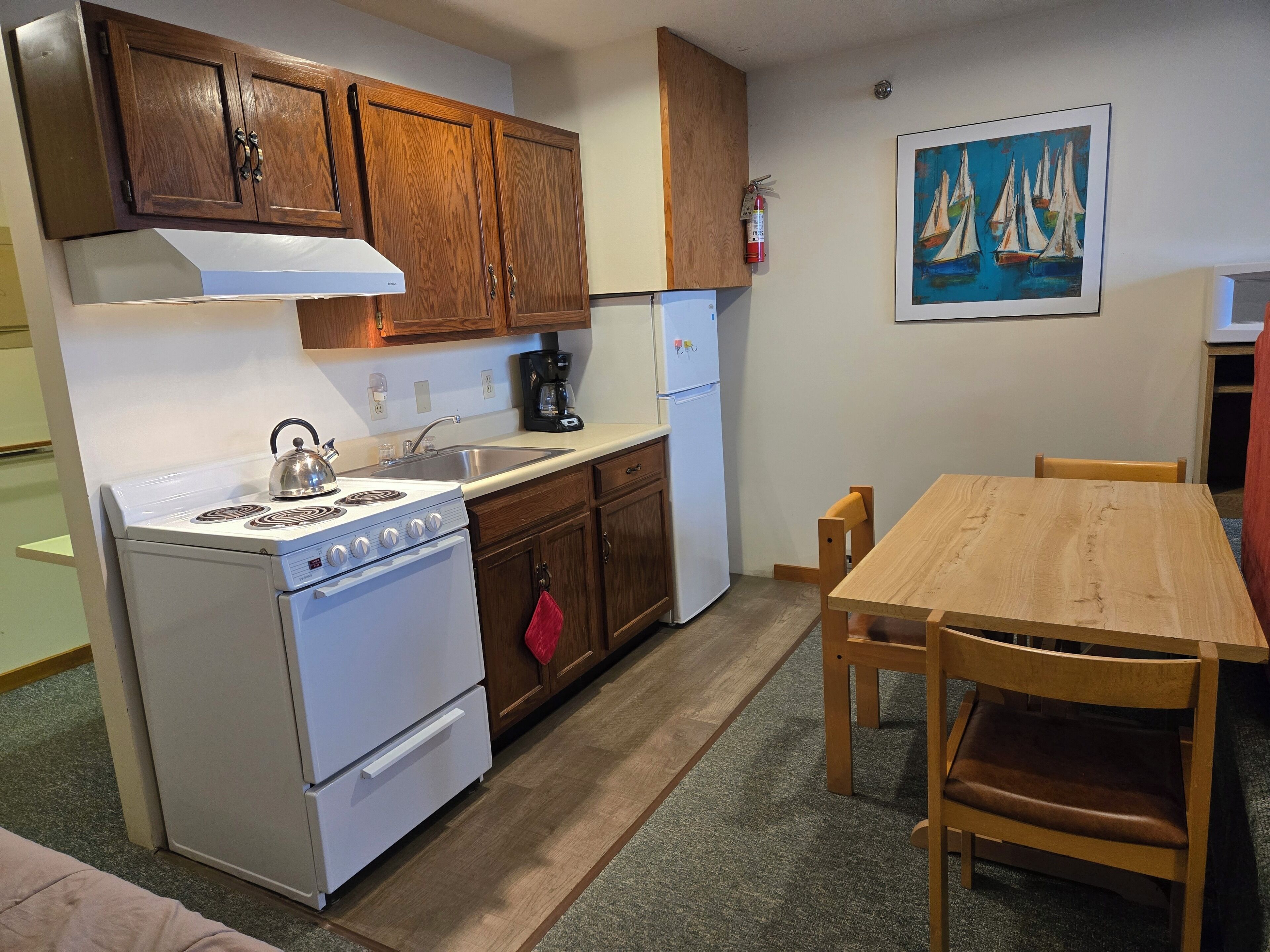 Una cocina compacta con armarios de madera, una estufa blanca, un lavamanos y una cafetera. Hay una mesa y sillas de madera, y un cuadro de veleros en la pared.