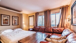 Quarto de hotel com cama, mesa de centro de madeira, sofá de couro e janela com cortinas.