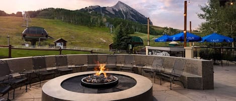 A circular outdoor seating area with a fire pit, surrounded by chairs and tables, overlooking a mountainous landscape.