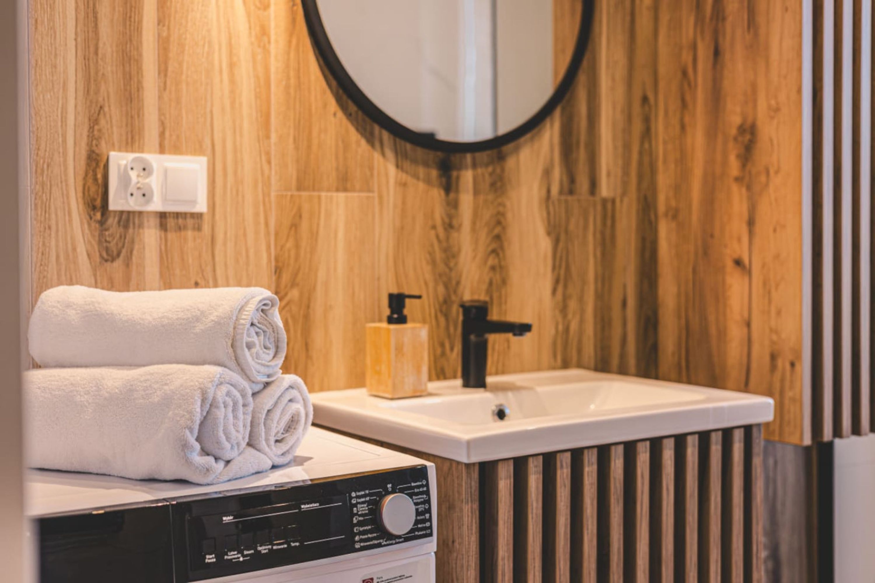 A bathroom with a wooden wall, a white sink, a black faucet, a soap dispenser, and a rolled white towel on a white surface.