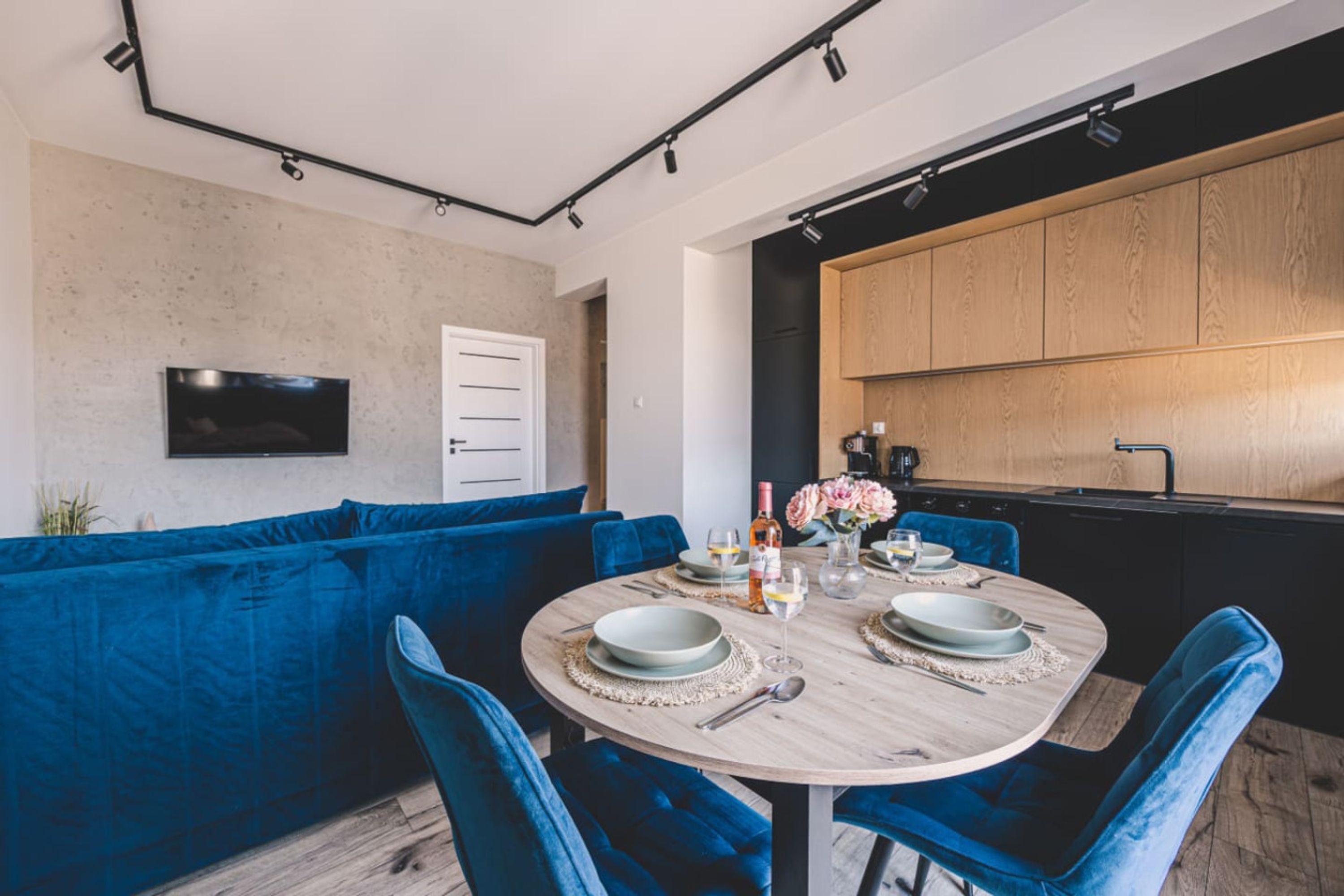 A modern dining area with a round table set for four, blue velvet chairs, and a kitchen with wooden cabinets in the background.