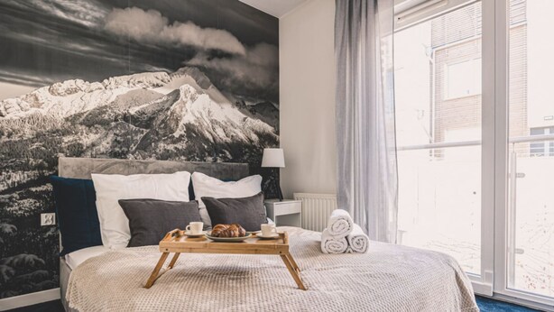 Ein Hotelzimmer mit Bett, einem Tablett mit Frühstücksgegenständen, einem Wandbild mit Bergmotiv und Blick ins Freie.