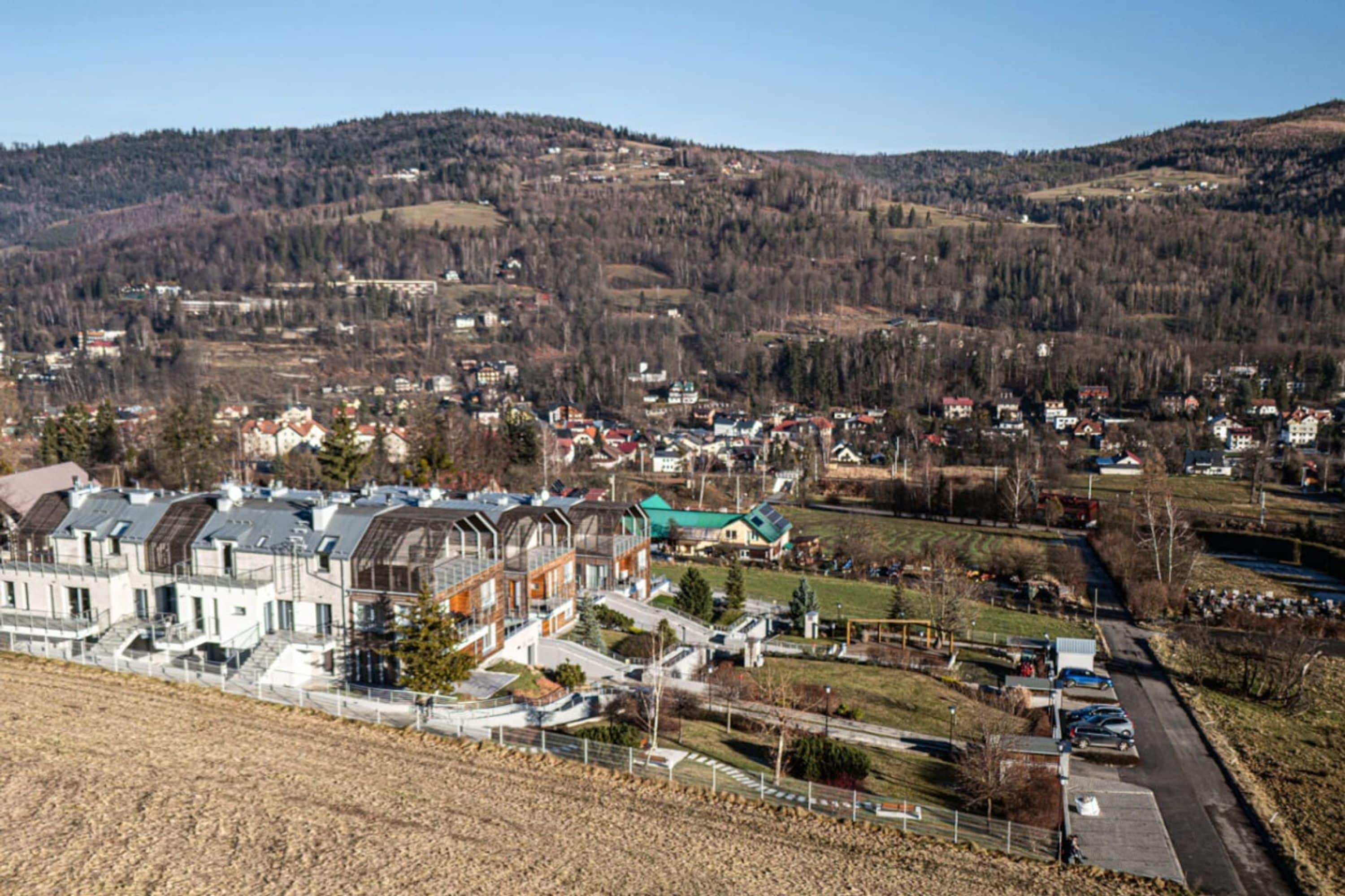 A residential area with modern buildings, a parking lot, and a hilly landscape in the background.