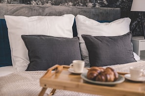 En seng med hvide og grå puder, et træbakke med croissanter og kopper, samt et natbord med en lampe.
