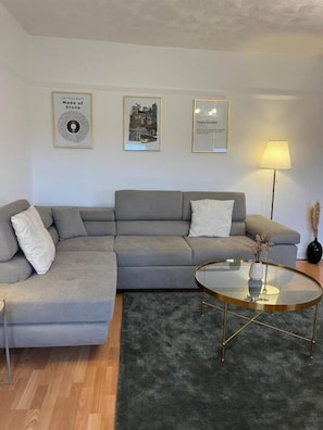 Ein modernes Wohnzimmer mit einem grauen Ecksofa, einem Glastisch und gerahmten Kunstwerken an der Wand.