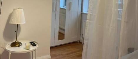 A room with a flat-screen TV mounted on the wall, a small table with a lamp, and a white curtain partially drawn.