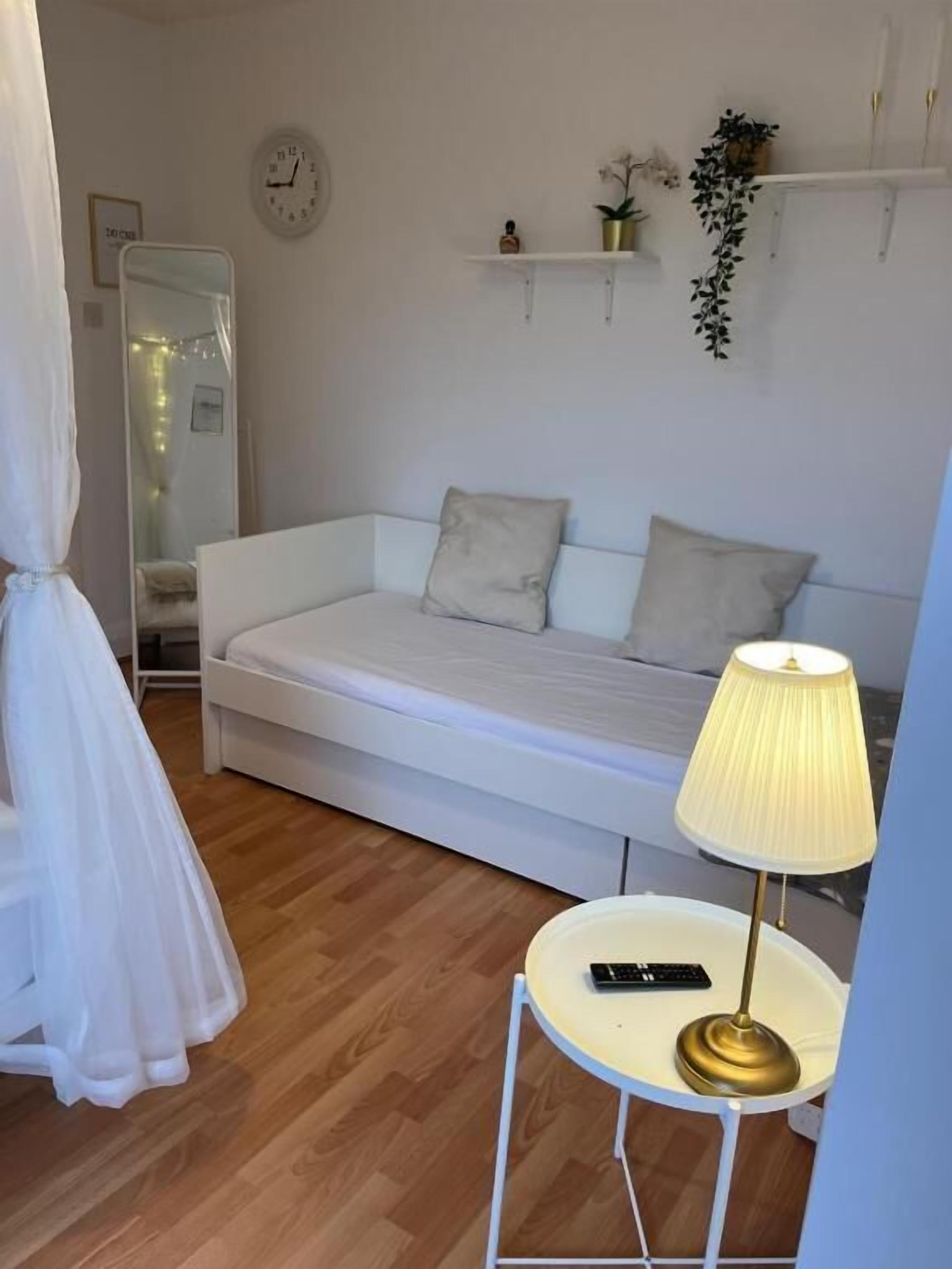 Ein weißes Bett mit Lampe, einem kleinen Tisch und einem Spiegel in einem Zimmer mit Holzboden.
