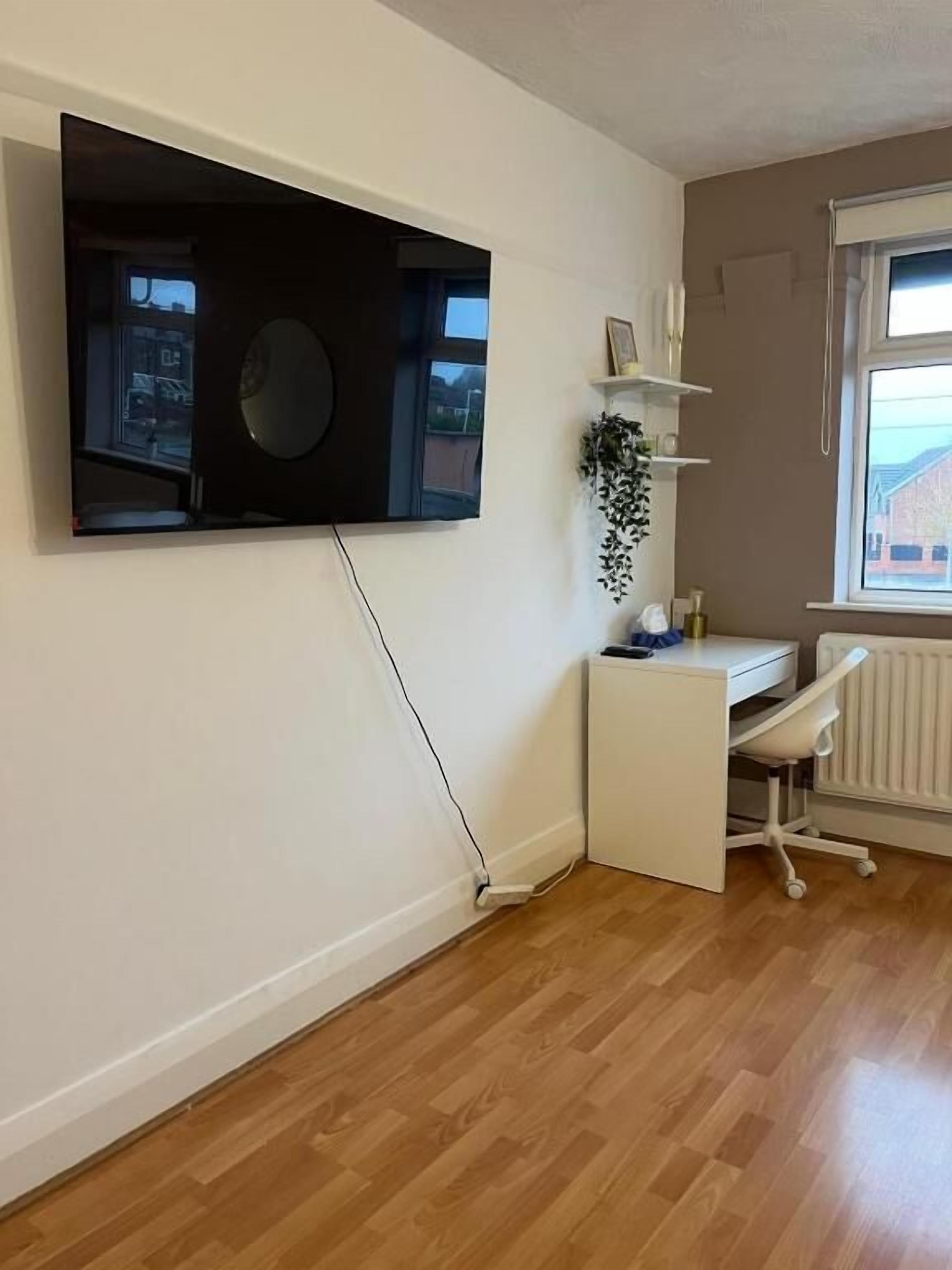 A room with a mounted TV, a white desk, a chair, a radiator, and a window with a view of buildings.