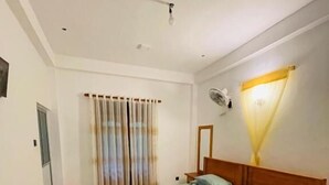 A bedroom with a bed, a bedside table, a mirror, a ceiling fan, and a window with curtains. - Rivers Edge Resort Sinharaja (Weddagala)