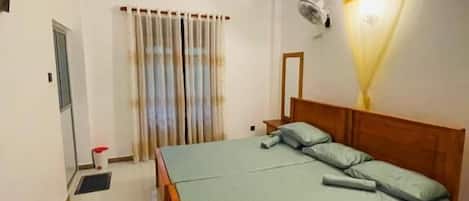 Een slaapkamer met een bed, nachtkastje, spiegel, plafondventilator en een raam met gordijnen.
