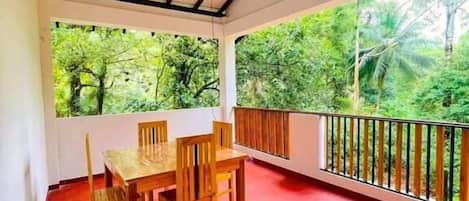 A covered outdoor area with a table and chairs, surrounded by a red floor and a railing overlooking a lush green landscape.