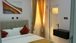 A bedroom with a bed, a bedside table, a wall-mounted air conditioner, a painting, a window with curtains, and a bathroom with a sink and mirror. - RBZ Apartments (Lagos)